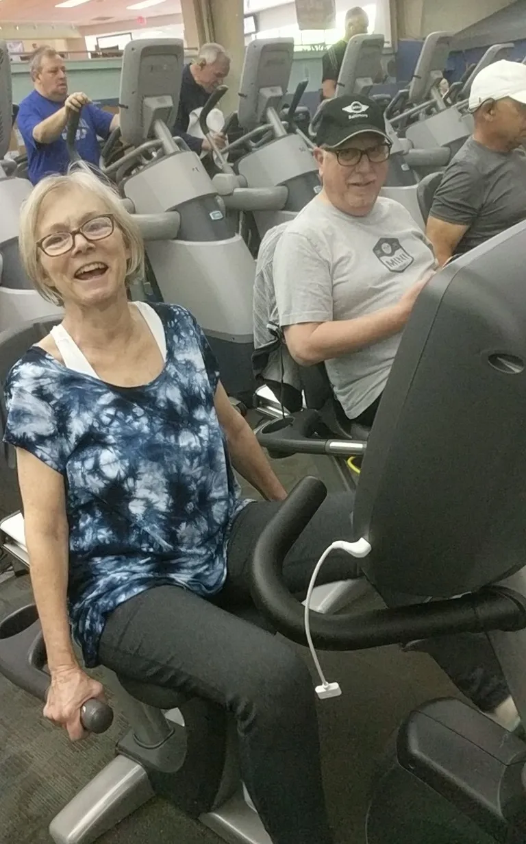 Suzanne and Ray laughing together on exercise bikes at the gym